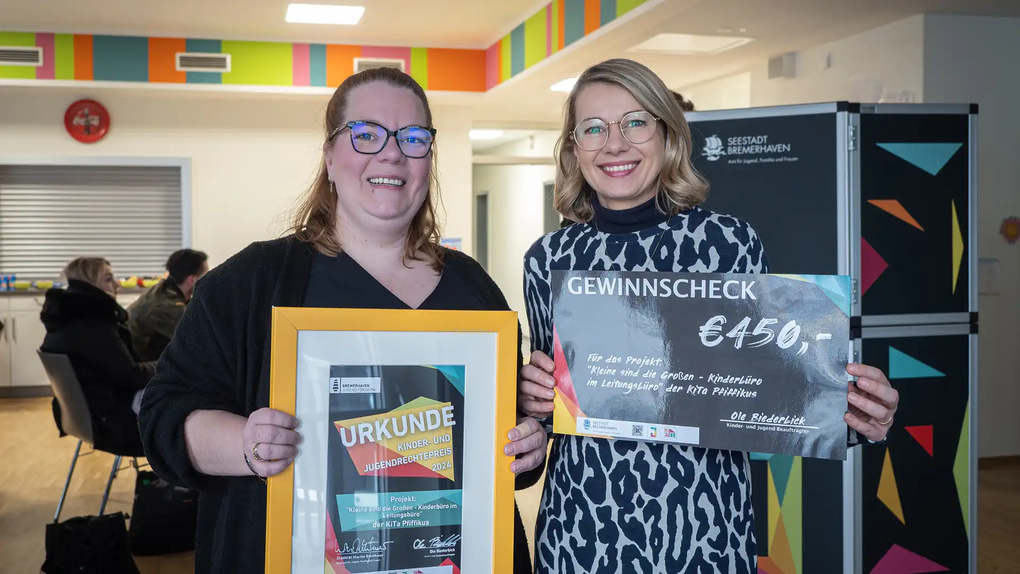 Das Foto zeigt zwei Frauen. Links Virginia Mibert. Rechts Cassandra Fee Berndt. Sie halten einen Gewinnscheck und eine Urkunde in ihren Händen.